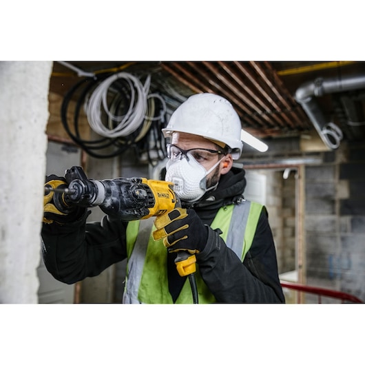 A person wearing a safety helmet, gloves, and a high-visibility vest is using a DEWALT power drill to work on a concrete wall in a construction environment. The face is blurred for privacy.