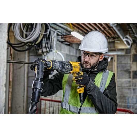 A person wearing a safety helmet, gloves, and a high-visibility vest is using a DEWALT D25134K rotary hammer drill on a construction site. The background shows exposed cables and piping in an unfinished building.