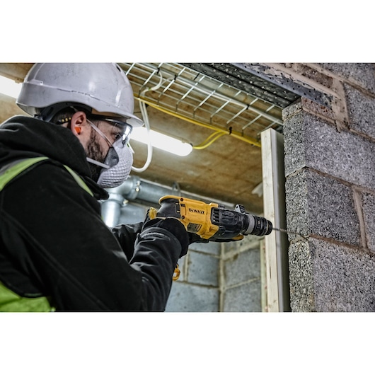 A worker wearing a safety helmet and gloves is using a DEWALT D25134K rotary hammer to drill into a concrete wall at a construction site.