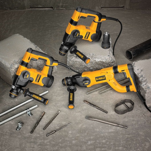 Three yellow and black DEWALT rotary hammer drills placed on pieces of concrete, with power cords, drill bits, and metal construction accessories scattered on the ground. The DEWALT logo is clearly visible on the tools.