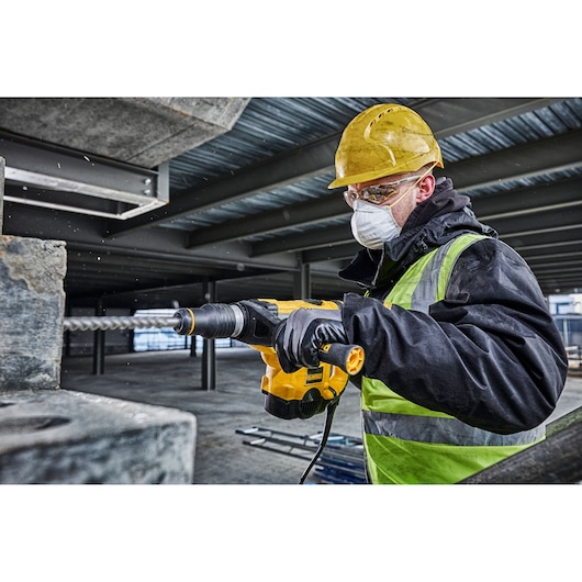 A person wearing a yellow hard hat, gloves, and a reflective safety vest is using a DEWALT power tool to drill into concrete in an industrial construction setting. The face is blurred for privacy.