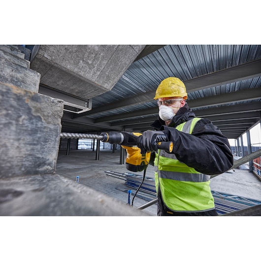 A construction worker wearing a yellow hard hat and high-visibility vest uses a DEWALT electric rotary hammer to drill into concrete at an industrial worksite.