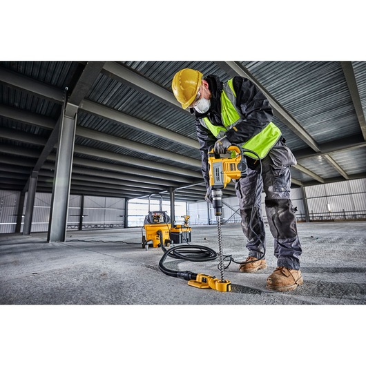 A worker in safety gear uses a DEWALT D25604K rotary hammer drill on a concrete floor inside an industrial building.