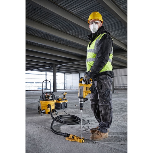 A worker wearing a yellow hard hat, reflective safety vest, and work boots is using a DEWALT rotary hammer drill (D25604K) connected to a dust extraction system on a concrete floor inside a large industrial building under construction.