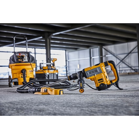 A DEWALT power tool set, including a yellow and black corded hammer drill with a dust extraction system attachment, displayed on a concrete floor in an industrial workspace. Additional DEWALT equipment is visible in the background.