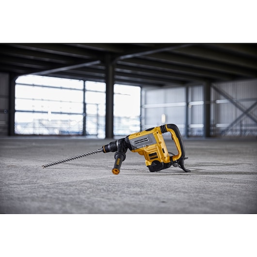 A DEWALT rotary hammer drill with a long drill bit, placed on the concrete floor of a large industrial building.