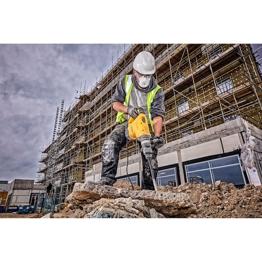 A construction worker wearing a safety helmet and high-visibility vest is using a yellow DEWALT demolition hammer to break concrete at a construction site. Scaffolding and an unfinished building are visible in the background.