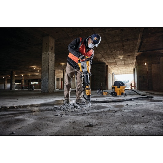 A construction worker wearing safety gear uses a DEWALT demolition hammer to break concrete in an unfinished building. A DEWALT dust extractor is visible in the background.