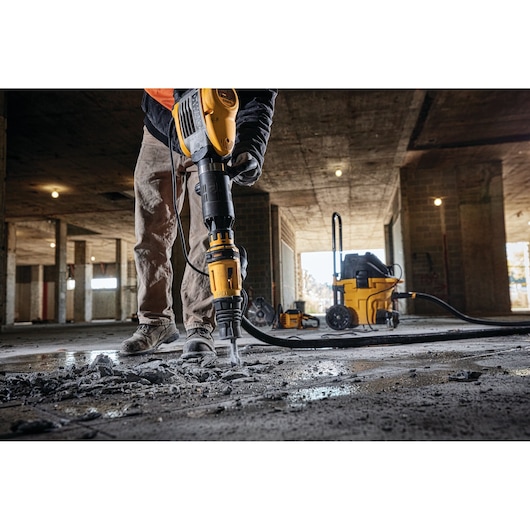 A person using a DEWALT D25951K demolition hammer to break concrete on a construction site, with debris scattered on the floor and additional DEWALT equipment visible in the background.