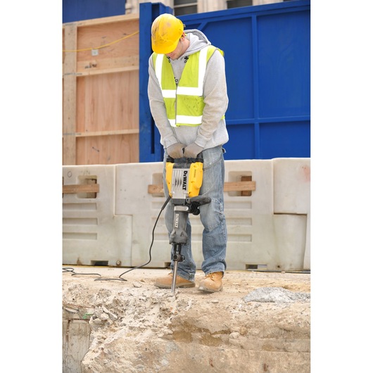 A person wearing a yellow hard hat, safety gloves, and a reflective safety vest is using a DEWALT electric jackhammer (model D25961K) to break concrete at an outdoor construction site.