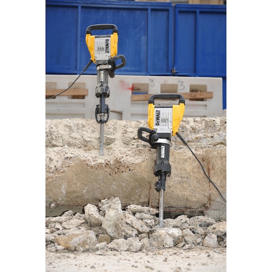 Two DEWALT electric demolition hammers are shown breaking up a large concrete slab, with rubble and broken pieces of concrete on the ground. The tools are positioned vertically and connected by cables, in an outdoor construction setting.
