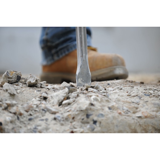 Close-up view of a construction worker's boot and a chisel breaking concrete on a worksite.