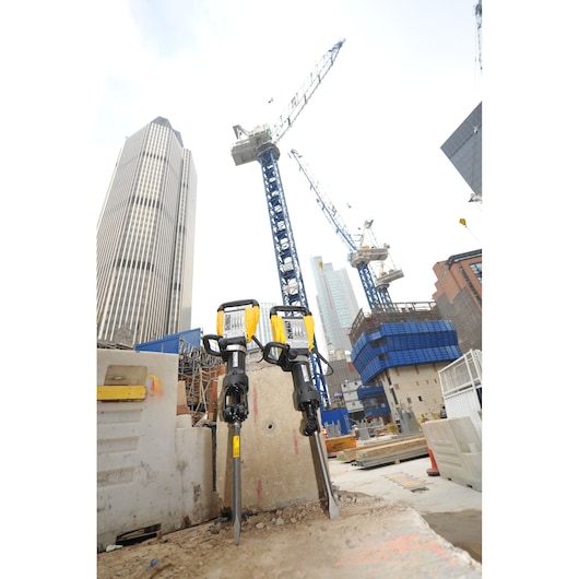 Two DEWALT demolition hammers placed on a concrete slab at a construction site, with cranes and tall buildings in the background.