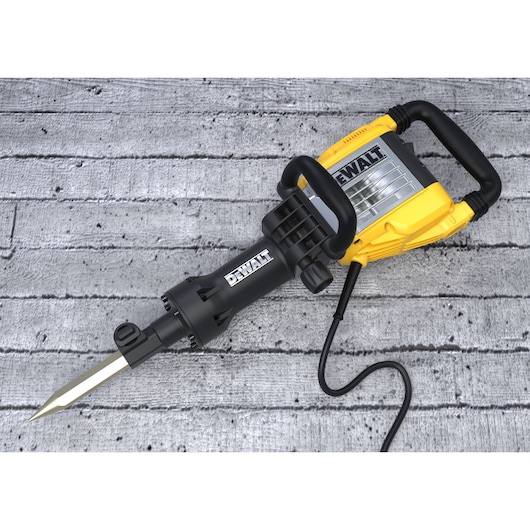 A DEWALT branded electric demolition hammer with a pointed chisel, featuring a yellow and black handle and casing, lying on a textured concrete surface.