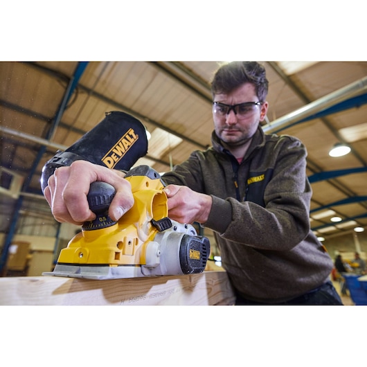 A person using a yellow DEWALT D26500 planer on a piece of wood in an indoor workshop. The person's face is blurred, and they are wearing a DEWALT-branded sweater.