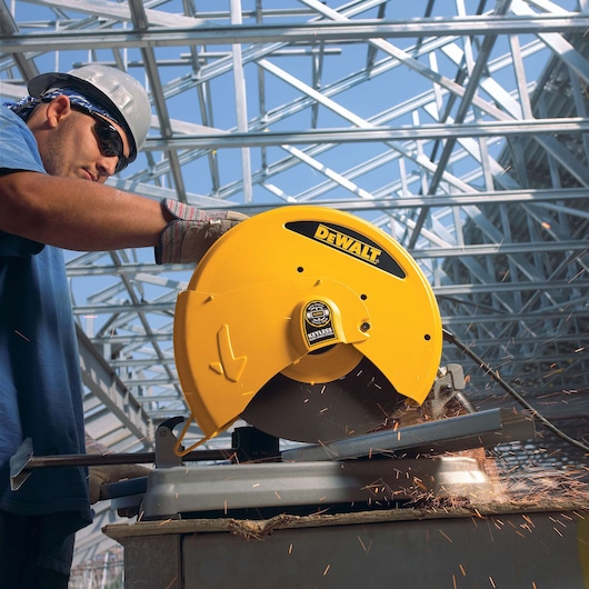 A person wearing work gloves and a hard hat operates a DEWALT yellow chop saw to cut metal, with sparks flying, inside a building with steel framework.
