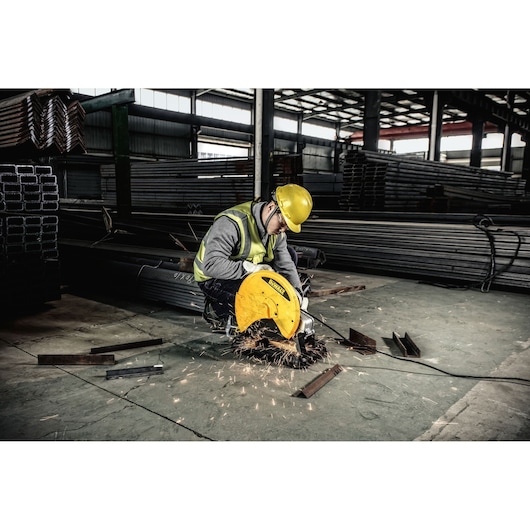 A person wearing a yellow safety helmet and high-visibility vest operates a DEWALT chop saw, cutting metal pieces in an industrial warehouse with metal beams stacked in the background.