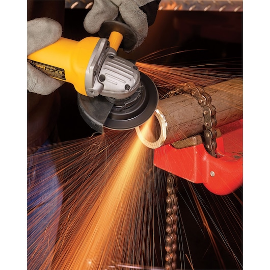 A close-up of a person using a yellow DEWALT angle grinder to cut through a metal pipe, with sparks flying from the cutting area. The pipe is secured in place with a chain vise.