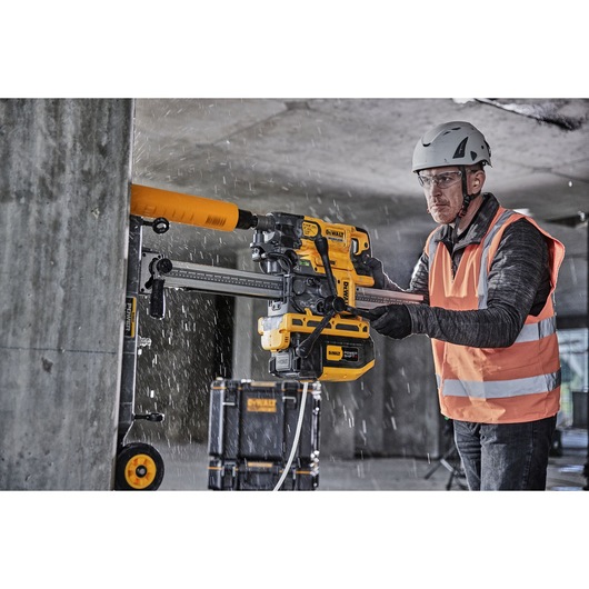 Worker uses a DEWALT drilling tool on a concrete wall; wearing safety gear and an orange vest.