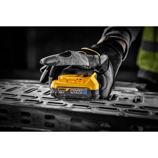 Gloved hand holds a DEWALT 18V POWERSTACK battery on a metal surface in a workshop setting.