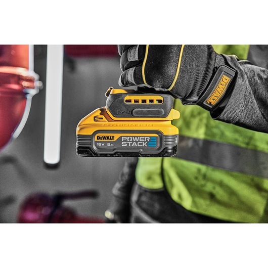 A gloved hand holds a DEWALT POWERSTACK 18V battery in a workshop setting, with safety gear visible.