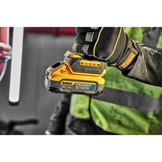 Gloved hand holding a DEWALT POWERSTACK 18V battery, close-up on the yellow and black product.
