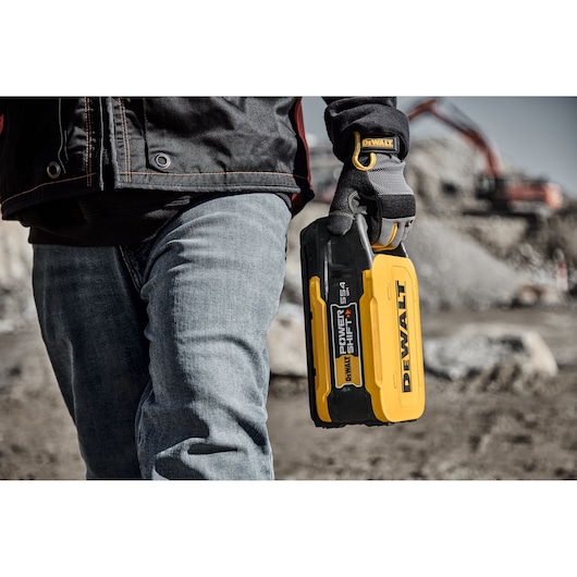 Person holding DEWALT DCBPS0554 battery outdoors at a construction site, wearing gloves and jeans.