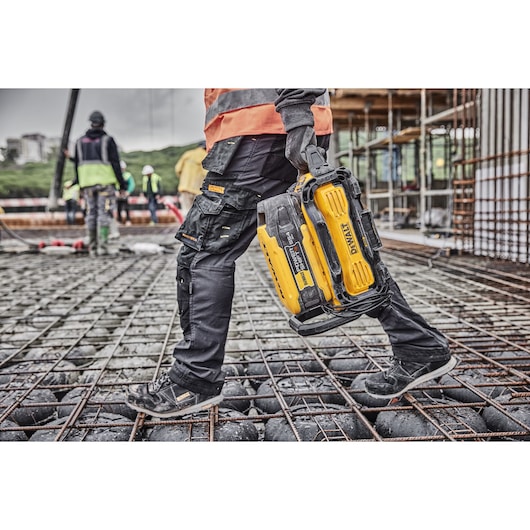Person carrying a DEWALT DCBPS0554 battery on a construction site, wearing safety gear and work boots.
