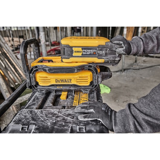DEWALT Power Shift battery being placed in a yellow DEWALT device at a construction site, handled by a gloved hand.