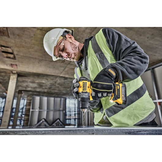 A construction worker wearing a safety helmet, high-visibility vest, and gloves is using a DEWALT DCD800E2T cordless drill to drill into a metal surface at an indoor construction site.