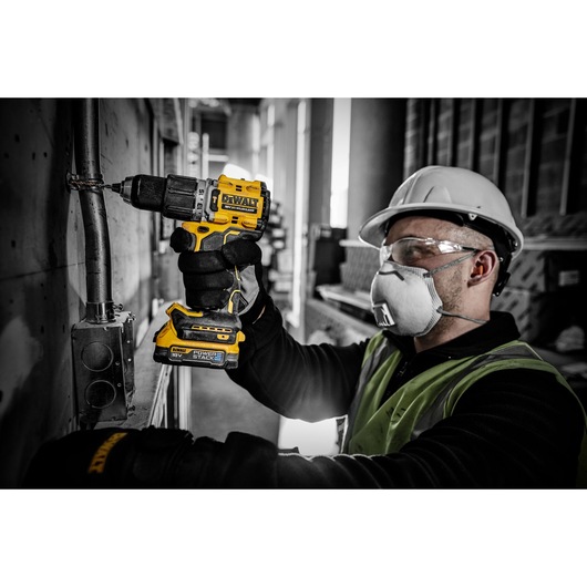 Worker uses DEWALT DCD805E2T cordless drill on a metal pipe. Wearing safety gear in an industrial setting.