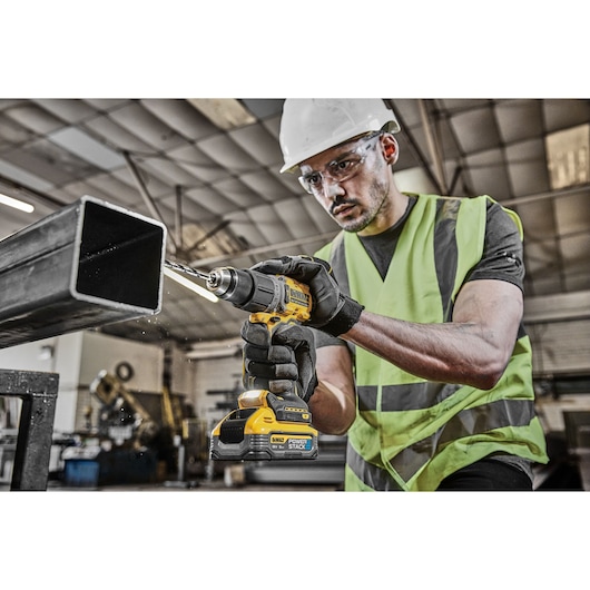 Worker using a DEWALT drill on a metal beam in an industrial setting, wearing safety gear and gloves.