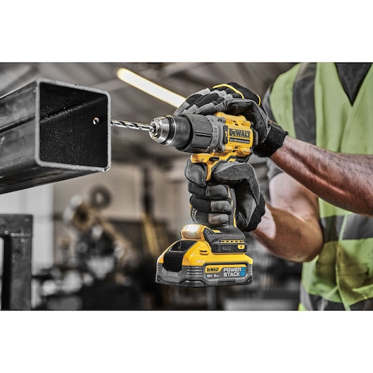 Gloved worker drills metal using a yellow DEWALT cordless drill in an industrial workshop.