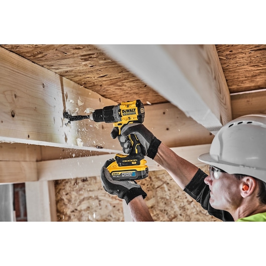 Person uses DEWALT cordless drill to make a hole in a wooden beam at a construction site, wood chips flying.