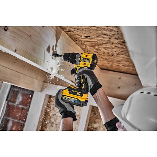 Person uses DEWALT cordless drill to bore into wood ceiling. Wood chips scatter. Worker wears gloves and helmet.