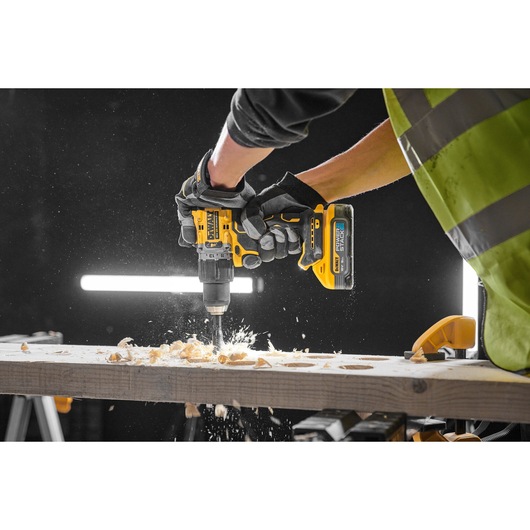 Gloved hands use a yellow DEWALT drill to work on a wooden plank, creating wood shavings and sparks.
