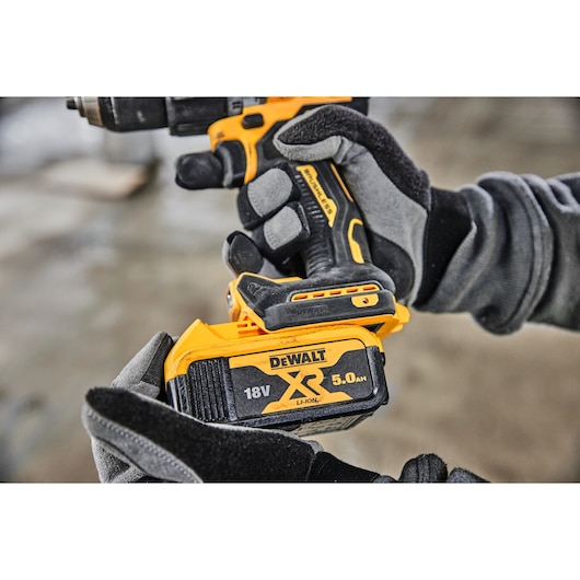 Close-up of a person wearing gloves holding a DEWALT power drill, focusing on the user inserting an 18V XR 5.0Ah Li-Ion battery into the tool.