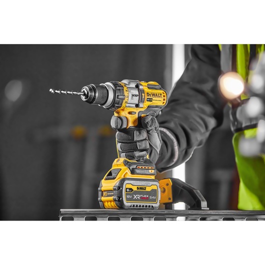 DEWALT DCD999T1 cordless drill with battery, held by gloved hand in a workshop setting.