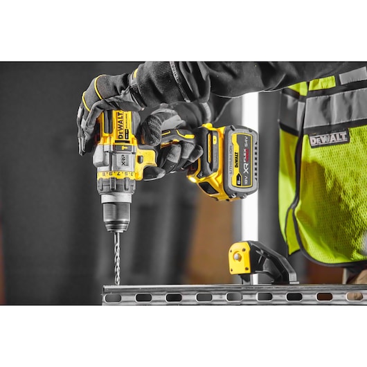 Gloved hand using DEWALT cordless drill DCD999T1 to drill into metal, construction vest visible.