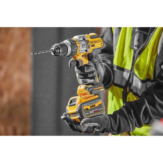 Person in work gear holding a DEWALT cordless drill with battery pack, indoors at a construction site.