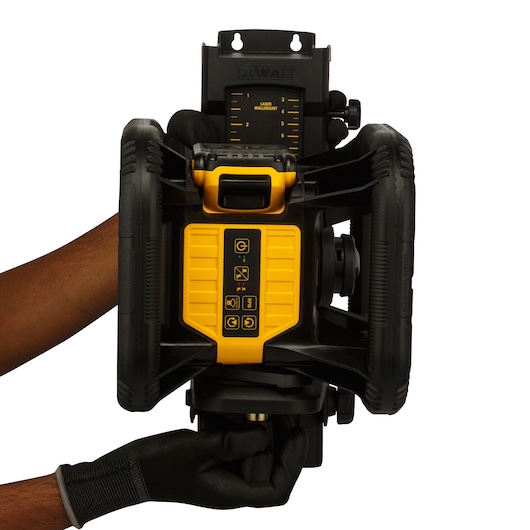A person wearing black gloves is adjusting a DEWALT rotary laser level, model DCE074D1R-QW. The yellow and black device is shown from the front, with control buttons visible and hands holding it securely.