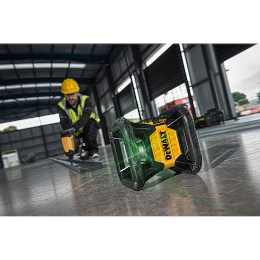 DEWALT laser level projects a green line on a construction site floor, worker in background with power tool.