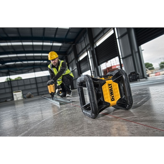 A DEWALT rotary laser level (SKU: DCE079D1R) is placed on the floor of an industrial construction site, projecting a red laser line. In the background, a construction worker wearing a yellow hard hat and high-visibility vest is operating equipment. The setting is a large indoor building with metal walls and open roller doors.