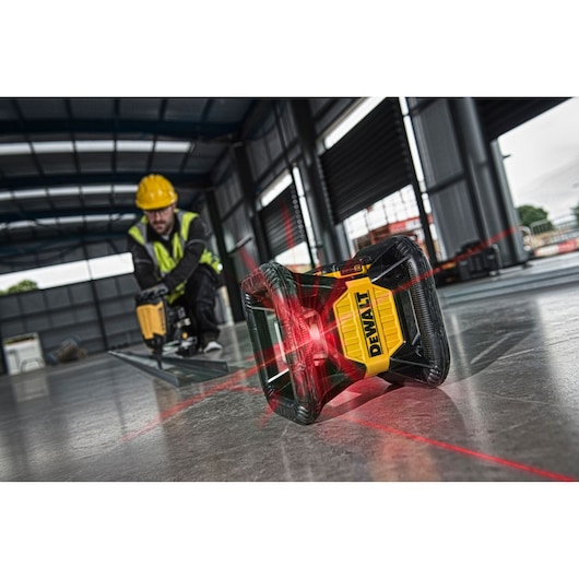 DEWALT red laser level projecting lines on a floor in an industrial building, worker in background using power tool.