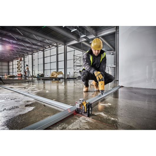 A construction worker using a Dewalt laser level (DCE089D1R) to align metal framing on a wet concrete floor in an industrial setting. The worker is wearing a yellow hard hat and operating a power drill while kneeling.