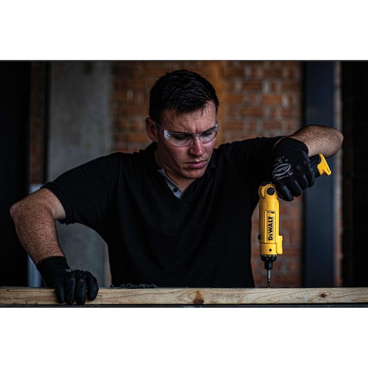 A person wearing black gloves is using a yellow DEWALT power tool to drill into a piece of wood. The background features a brick wall, and the person's face is obscured.