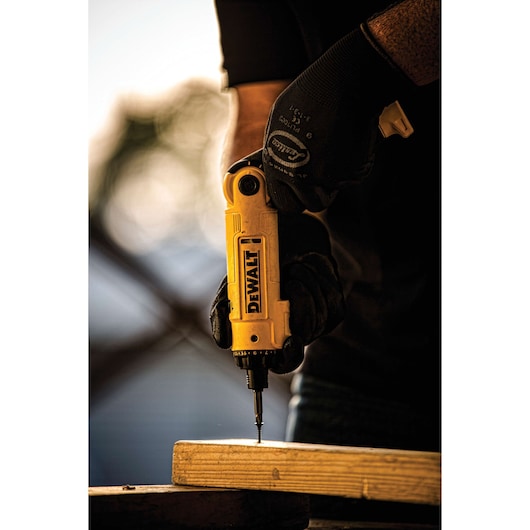 Close-up of a person wearing gloves using a yellow DEWALT DCF008 power screwdriver to drill a screw into a piece of wood.