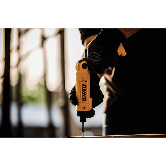 A person wearing black gloves is using a yellow DEWALT cordless screwdriver to screw into a surface, with a blurred industrial background.