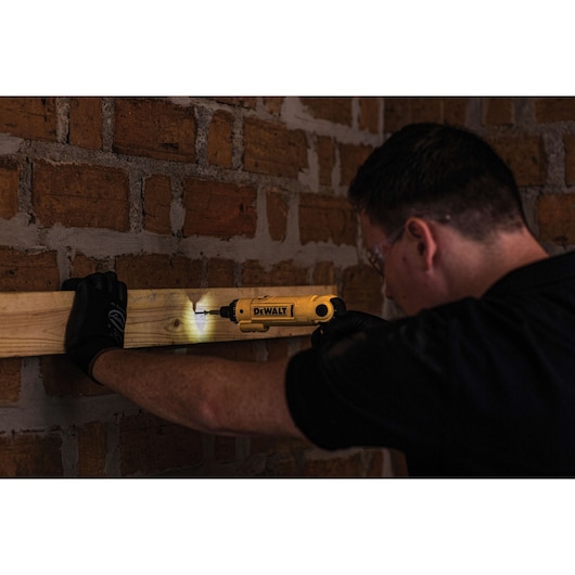 A person wearing gloves is using a DEWALT cordless screwdriver (model DCF008) to fasten a screw into a piece of wood attached to a brick wall. The tool's built-in LED light is illuminating the work area.
