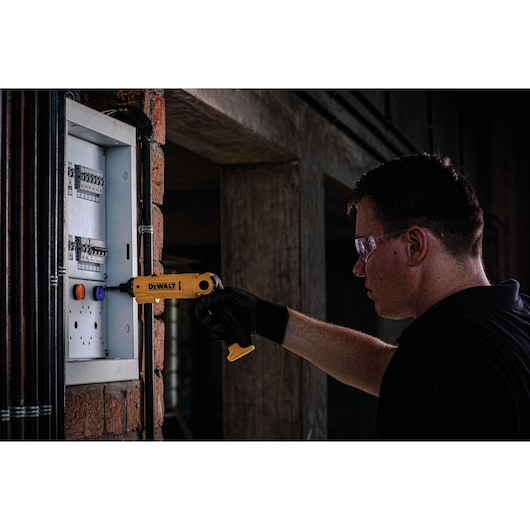A person wearing black gloves is using a yellow DEWALT tool to work on an electrical panel mounted on a brick wall. The person's face is obscured.